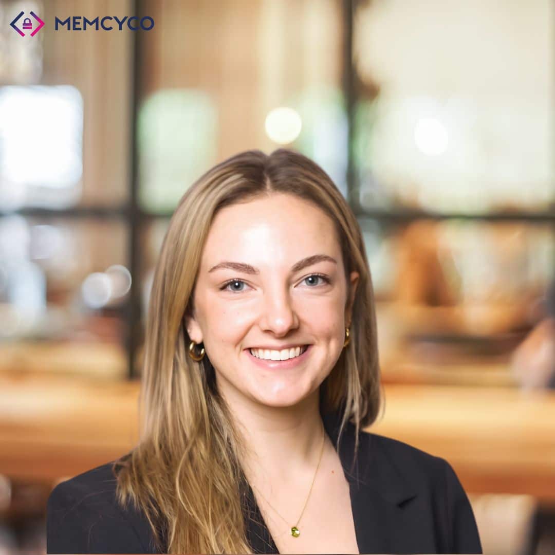 A woman with long blonde hair and a black blazer smiles at the camera; the background is a bright, modern office, highlighting the professional atmosphere. The MEMCYCO logo is in the top left corner, reflecting Memcyco at Global Anti-Scam Summit America 2026.