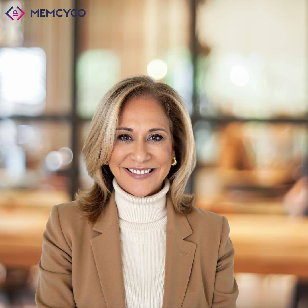 A woman wearing a tan blazer and white turtleneck smiles in a modern office with a blurred background, capturing the professional energy of Memcyco at Datos Insights FinCrime Forum 2026. The MEMCYCO logo appears in the top left corner.